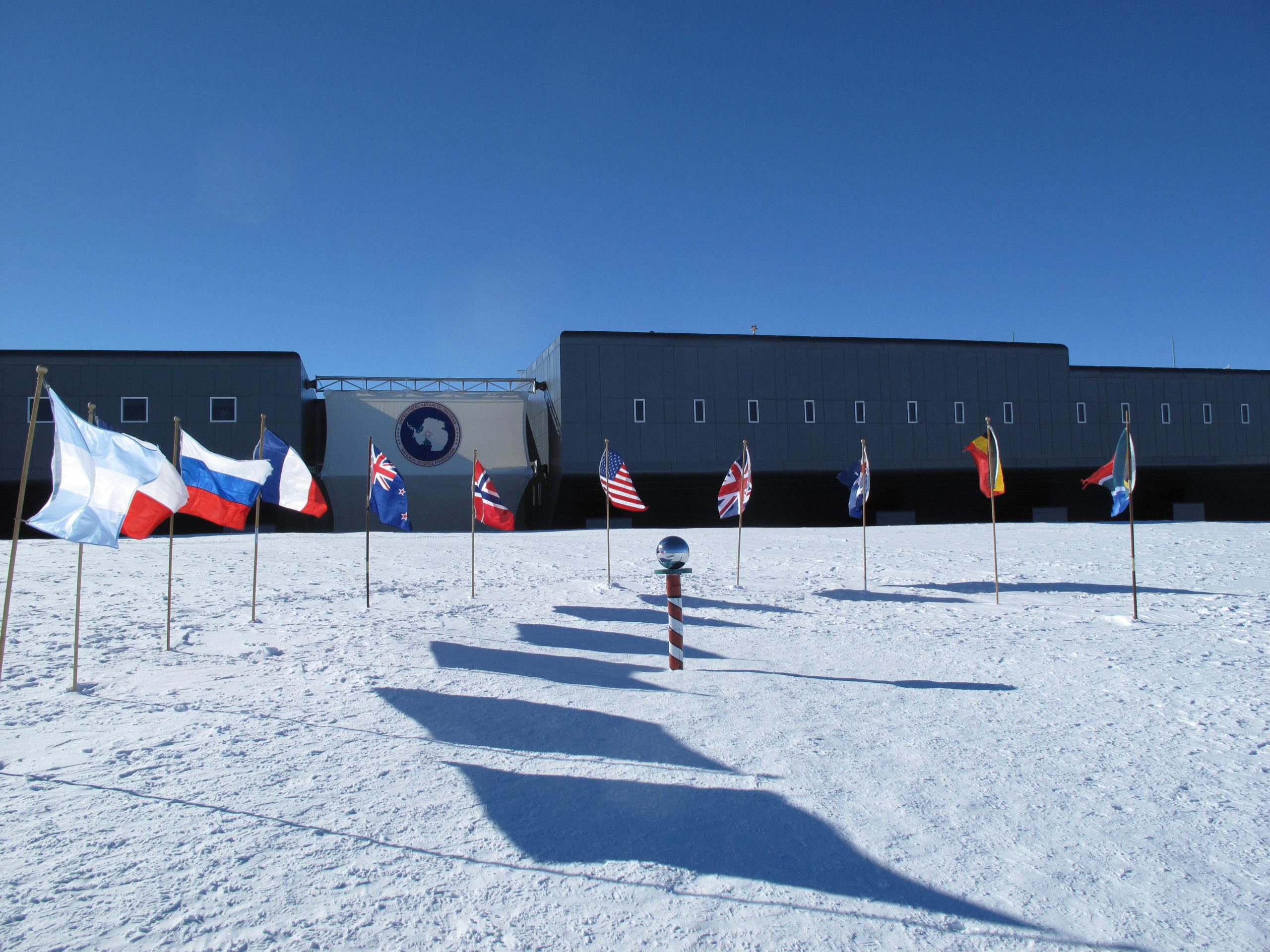 A group of flags with the amundsen scott south pole station in the background.