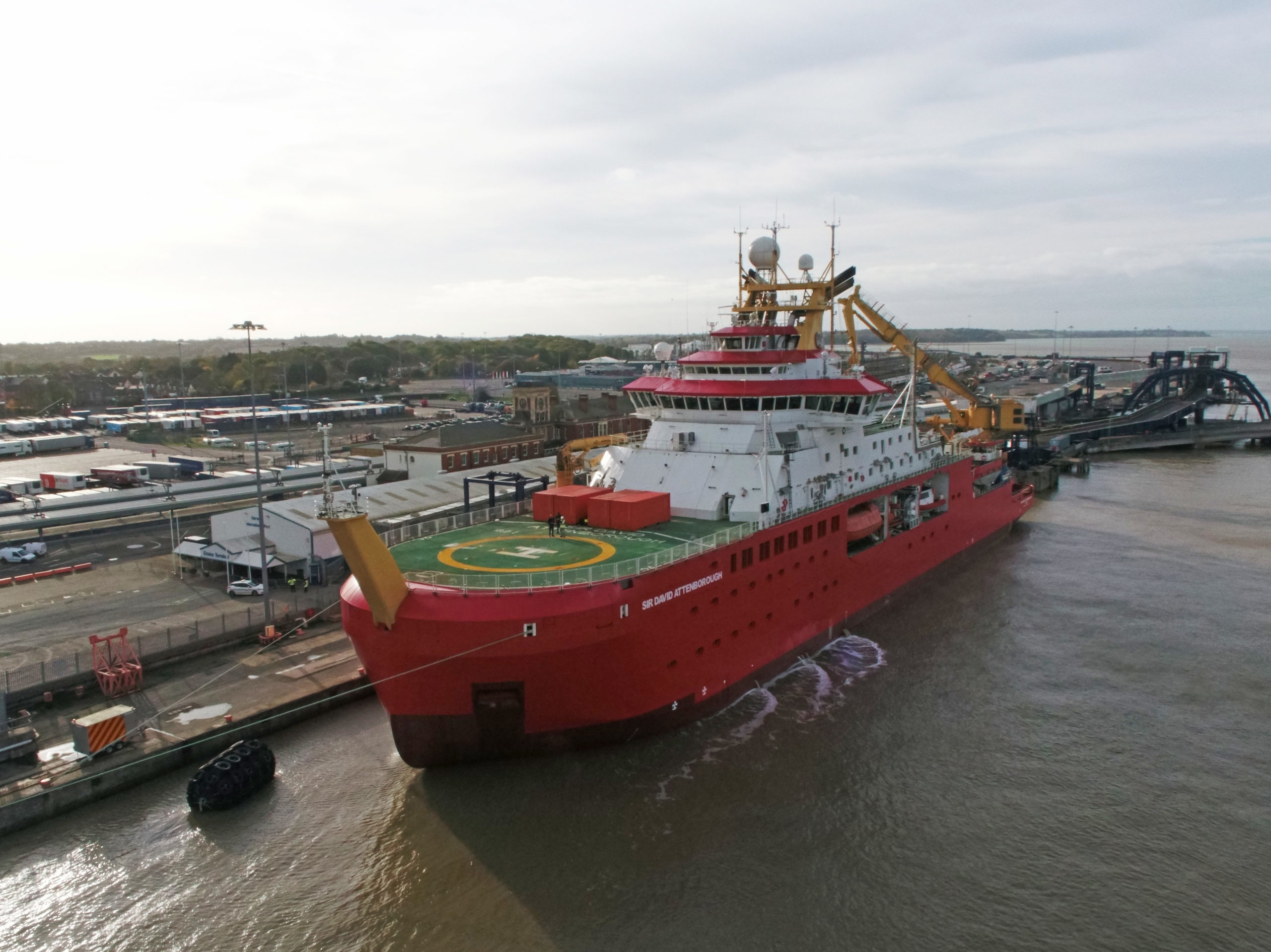 A picture of RRS Sir David Attenborough from the bow of the ship, next to the port. The ship is red and there is a green helideck visible, with a large white letter H on it. There are two large red containers on the deck. The superstructure is white, and you can see the ship's biggest crane which is yellow.