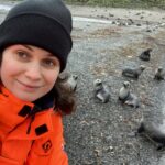 A smiling woman wearing a woolly hat on a rocky beach with some seals