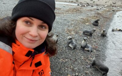 A smiling woman wearing a woolly hat on a rocky beach with some seals