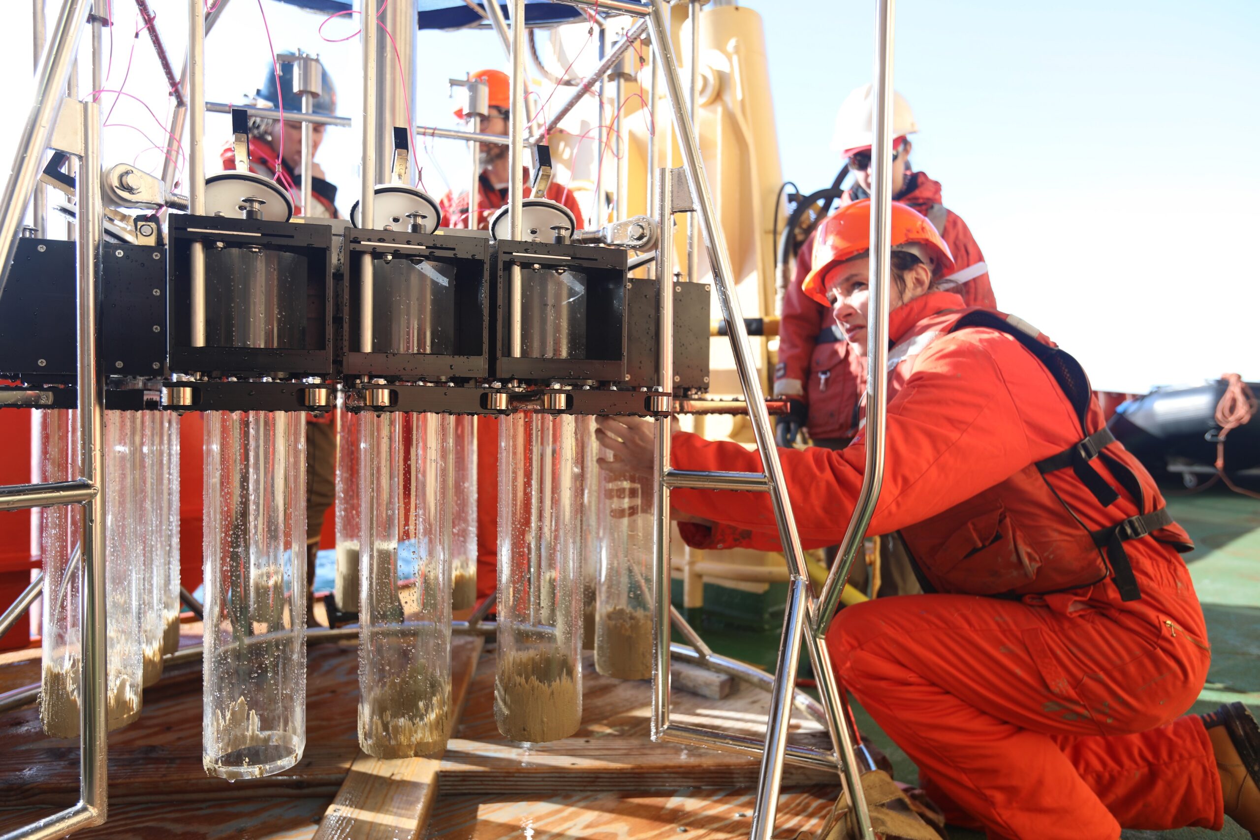 A woman in a boat suit crouches down and touches a piece of science equipment on the deck of a ship