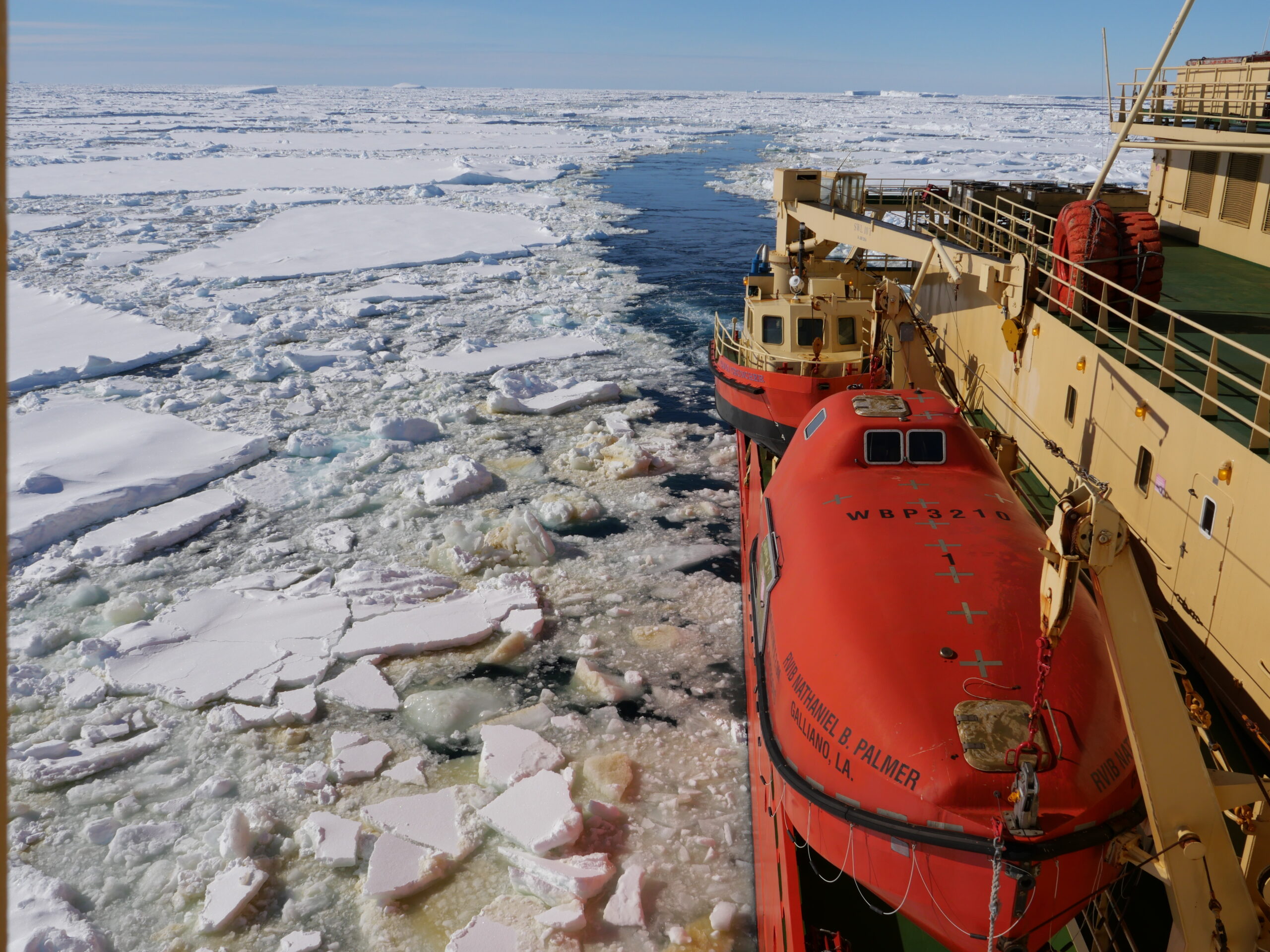 The side of a ship in sea ice. A red lifeboat is visible.