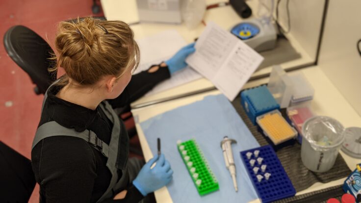 A young woman, her hair tied up in a bun, is seen from above sitting at a desk, which holds a variety of scientific equipment (e.g. beakers, test tubes, sample racks). She is wearing blue gloves, and with one hand holds a sample of anemone DNA. Facing away from the camera, she is referring to a piece of paper in her other hand, which allows her to tell if the sample she is holding matches pre-existing genetic data, confirming whether the team have found a new species of mollusc