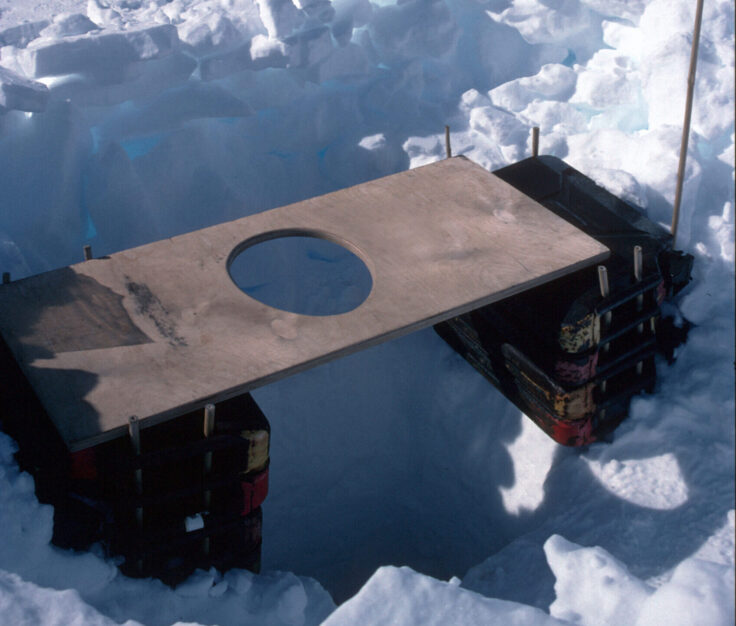 A wooden toilet seat on top of a hole dug into snow. 