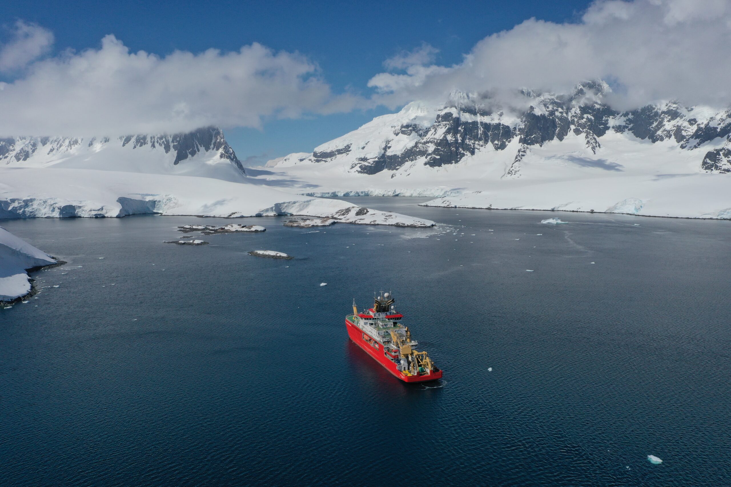 A red ship sailing through a flat ocean, with snow covered mountains behind