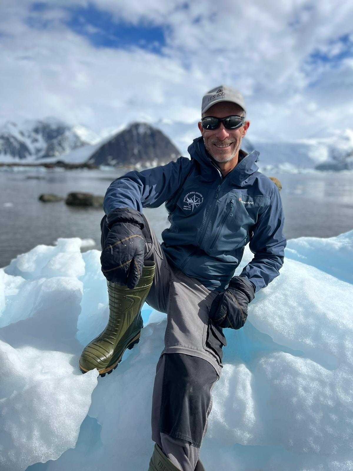 A smiling man wearing a baseball cap and sunglasses sitting on top of a block of ice with sea and ice capped mountains behind him against a cloudy sky.